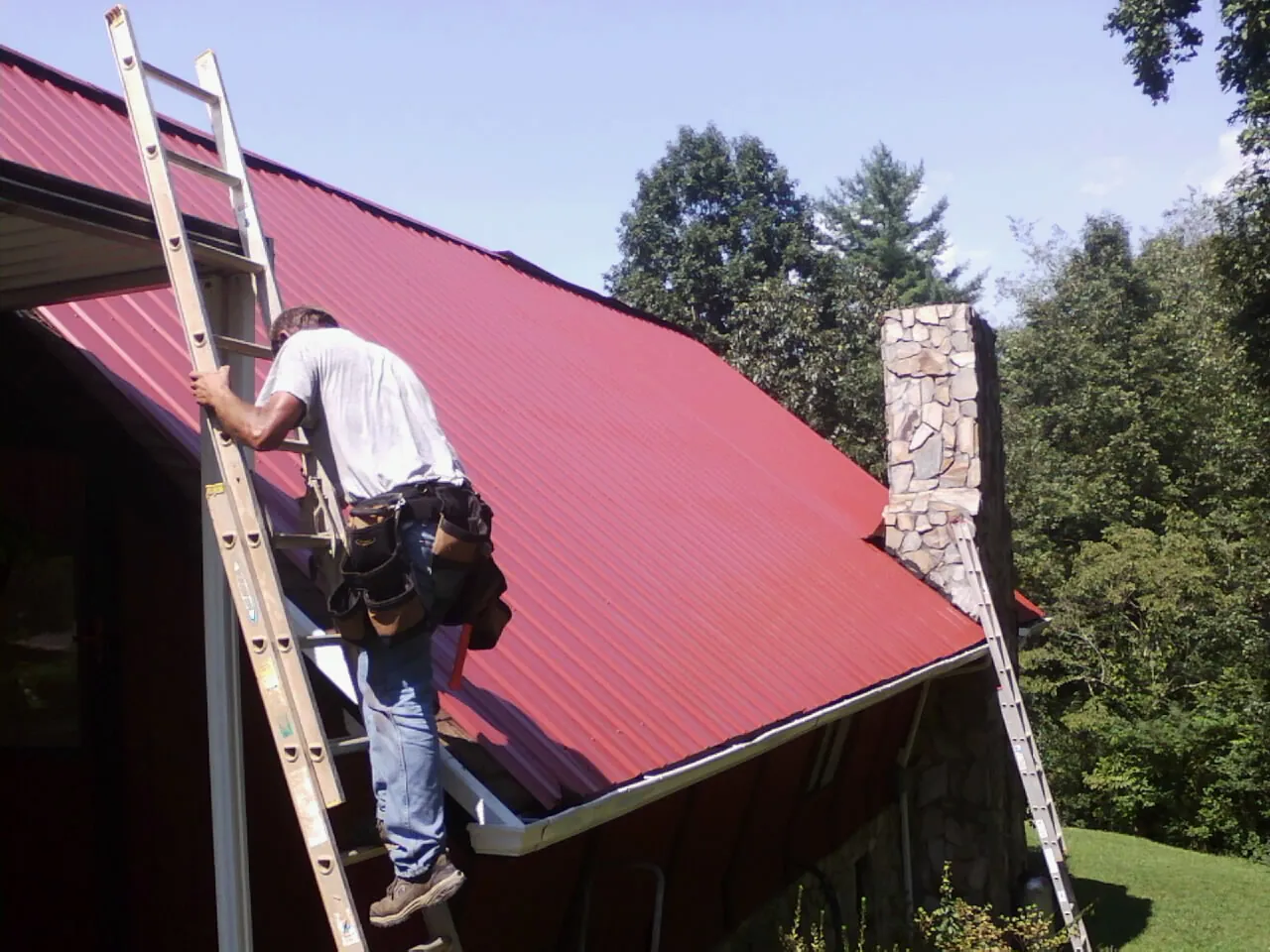 Roofer on a ladder beside a red metal roof and stone chimney — Soffit & Fascia Repair by Roofing Brothers in Adair